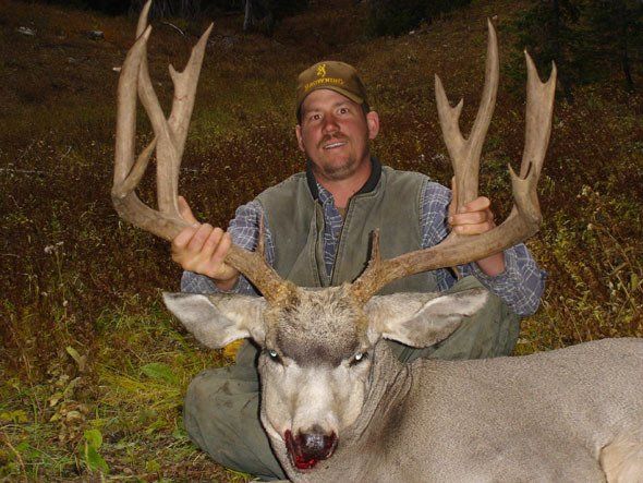 Man holding large deer antlers, deer in foreground. Brown hat, green vest, outdoors.