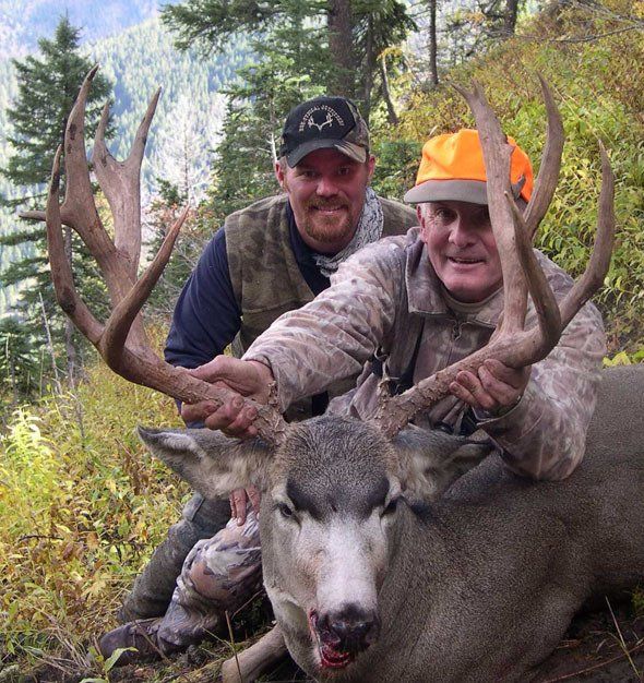 Two men posing with a large deer they hunted in a wooded area, smiling, with antlers.