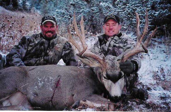 Two men in camo with a large buck, snowy field backdrop. Men smiling, posing with antlers.
