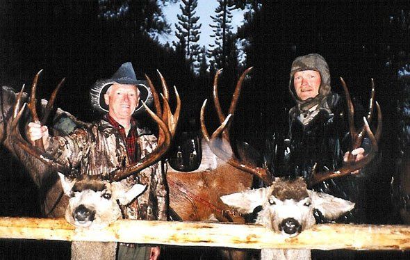 Two men pose with large deer antlers. They are outside at dusk. The men are smiling.