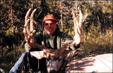 Man in orange cap and green hoodie with large deer antlers in a wooded area.
