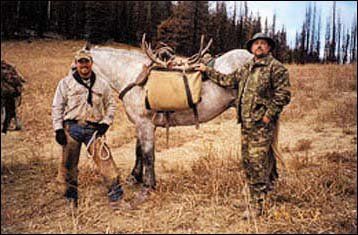 Two men with a saddled horse in a field; one man holds reins, the other poses with a sack and antler.