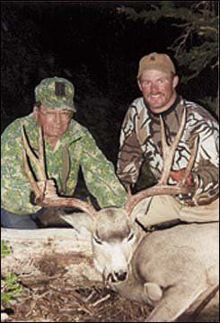 Two men in camo with a harvested deer; antlers prominent. Outdoor setting.