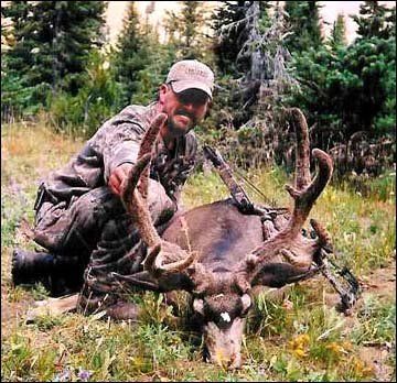 Hunter kneels next to a deer with large antlers in a forest. Man wears camo. Deer has white markings.