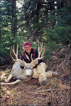 Hunter kneels beside a large, pale buck in a forest, holding the deer's antlers.