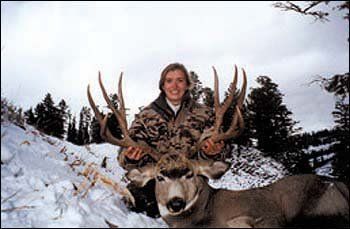 Woman in camo holds large deer antlers, snow-covered landscape.
