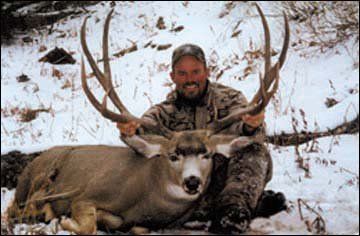 Man kneeling with large antlered deer in snow. He is smiling.