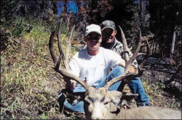 Two men kneeling behind a large buck with impressive antlers in a wooded area.