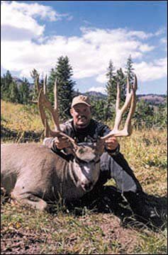 Man holding massive deer antlers, posed beside the harvested deer in a grassy outdoor setting with trees.