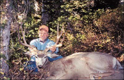 Man kneels beside a dead buck in a forest, holding its antlers.