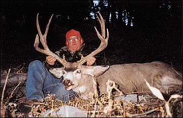 Hunter in a red hat holds a large buck with impressive antlers, sitting in brush.