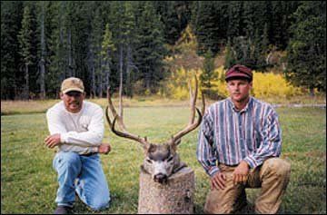 Two men pose with a large deer head trophy outdoors on a stump.
