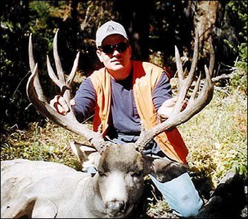 Man in sunglasses holds the large antlers of a dead deer. He's wearing an orange vest and is outdoors.