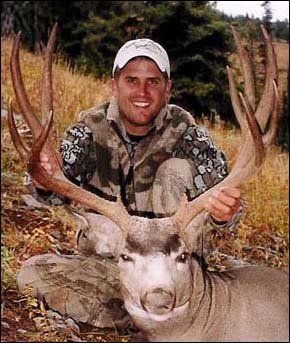 Man in camo holds large deer antlers, smiling in outdoor setting.