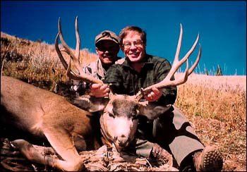 Two men posing with a deer they shot, outdoors. Sunny, blue sky.