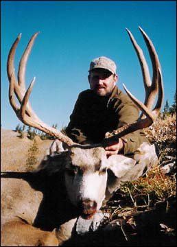 Man kneeling behind a large mule deer, sky blue, field.