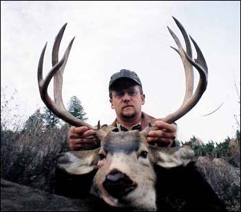 Man holding up the head of a deer with large antlers. Outdoors, brown hues, overcast sky.