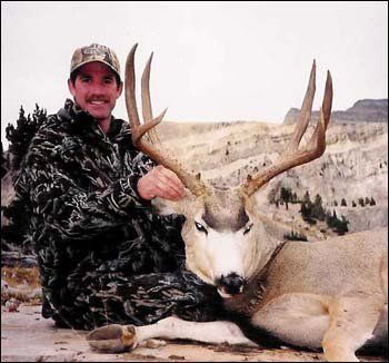 Man in camouflage kneels next to a large deer with impressive antlers, mountain backdrop.