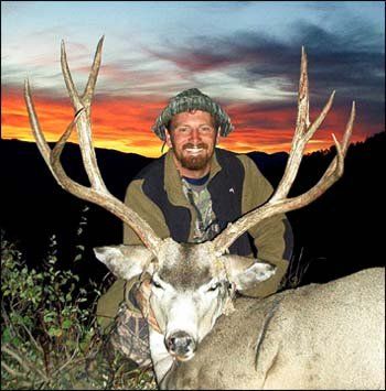 Man smiles, posing with a large deer he hunted at dusk with colorful sky.
