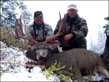 Two men pose with a large buck deer in a snowy forest.