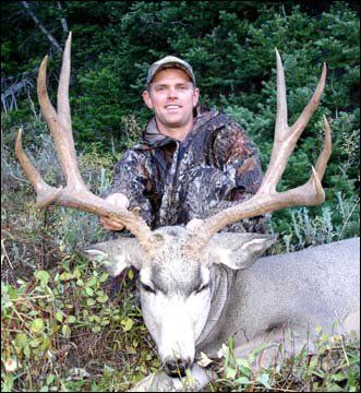 Man in camouflage holds a large buck with massive antlers, smiling in a forest setting.