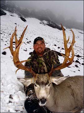 Man in camo holding deer with large antlers in snowy mountains.