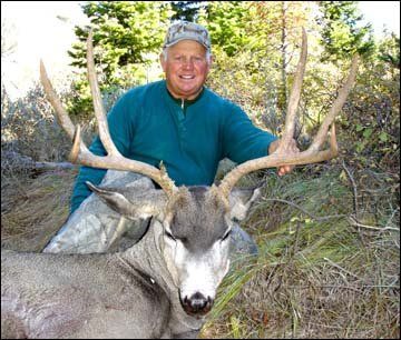 Man in green shirt smiles next to large buck with large antlers outdoors.