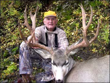 Hunter in camouflage crouches beside a large deer with impressive antlers, both are outdoors in a natural setting.