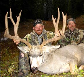 Two hunters with a large mule deer buck, outdoors, holding its impressive antlers; both are smiling.