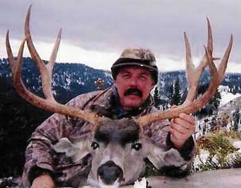 Man holding a large mule deer buck with impressive antlers outdoors in a mountainous setting.