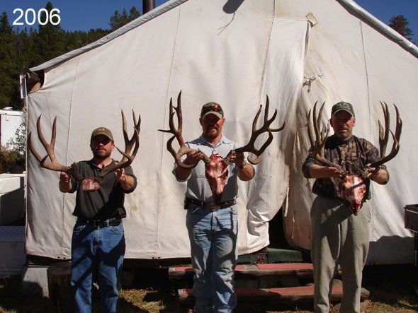 Three men holding large deer antlers in front of a white tent. 2006 is written in top left corner.