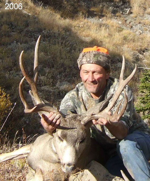 Man kneeling, holding deer antlers in a mountain setting. Deer is laying down. Man is wearing camouflage and orange cap.