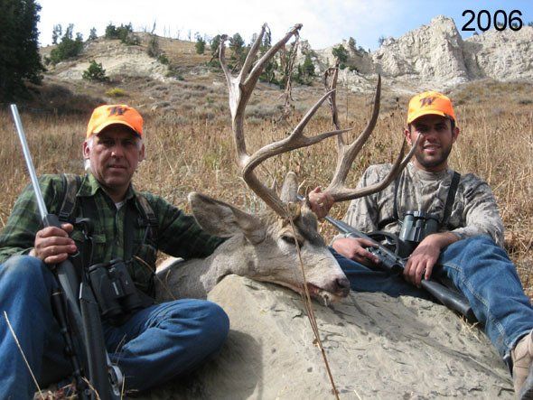 Two men in orange hats with a dead deer. One holds a rifle, the other rests it. The background has a rocky hillside.