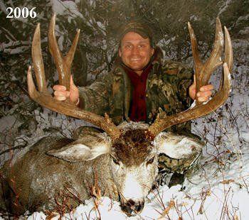 Man in camouflage holds up large deer antlers, smiling in the snowy forest.
