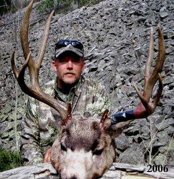 Man in camo with large deer head, antlers, in front of rock background; blue and white tape on the antlers; 2006.