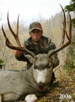 Hunter in camouflage with large mule deer buck; outdoors, fall foliage. Buck has large antlers.