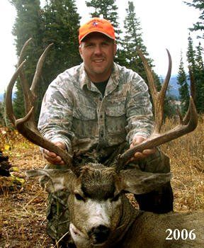 Man kneels, holding large deer antlers, with a blurred mountain backdrop and 