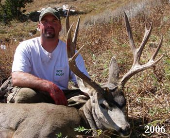 Man kneeling with a large buck, possibly after a hunt. The man smiles and the buck lays in grass. Year: 2006.