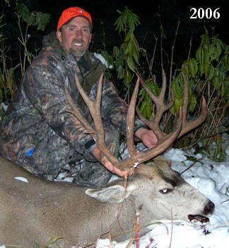 Hunter kneeling with a large buck in the snow; hunter wears camo and an orange hat, 2006.
