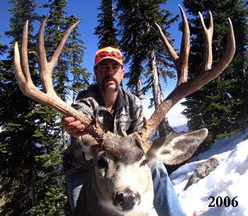 Hunter holding large buck antlers in a snowy forest; the deer's head is visible, with a blue sky in the background.