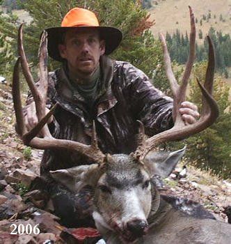 Man in hunting gear with a large deer he killed, holding up its antlers. He wears an orange hat and smiles.