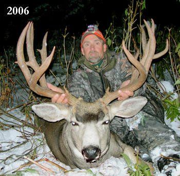 Hunter in camouflage holds a large buck with antlers in a snowy, wooded area.