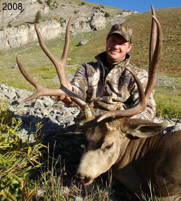 Hunter in camouflage holding large mule deer antlers in a mountain setting. Smiling, with 