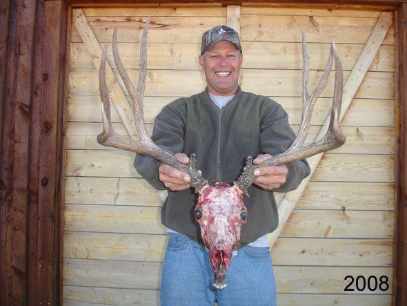 Man holding large deer skull with antlers, smiling. Wooden wall background, 2008.