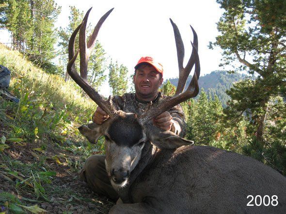 Man kneeling, holding large buck with impressive antlers in a forest setting; the man wears camouflage.