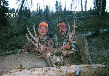 Two men in hunting gear pose with a large buck in a wooded area; the year 2008 is in the corner.