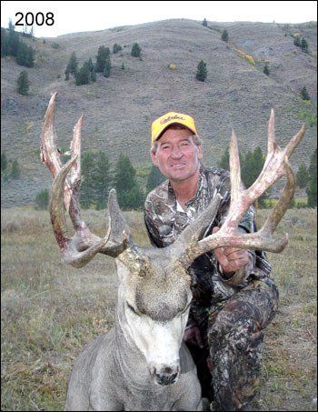 Hunter kneels next to a large-antlered deer in a grassy field. He smiles, wearing camouflage and a yellow hat.