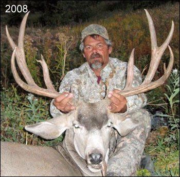 Man in camouflage holding a large deer's antlers. The deer lies in the grass. Text 