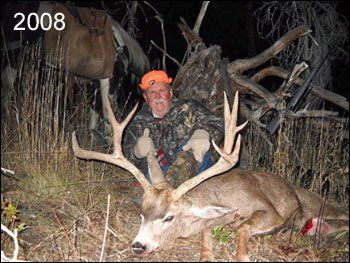 Hunter with a large buck, 2008. Man in orange hat and camo giving thumbs up. Buck is lying on ground.