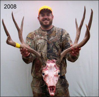 Man in camouflage holds up a deer skull with large antlers; wearing a bright orange hat, smiling. 